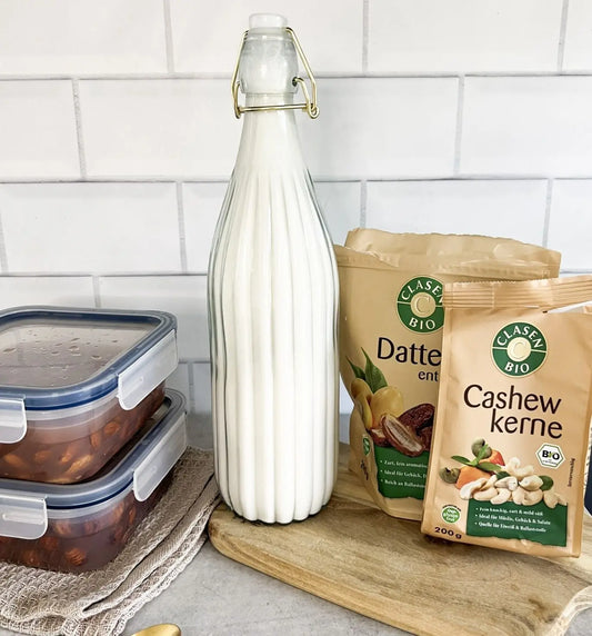 Hausgemachte Nussmilch aus Mandeln, Cashews und Haselnüssen in einem Glasbehälter auf einem Holzbrett, daneben Cashewkerne, eine Datteln-Verpackung und zwei Tupperdosen mit über Nacht eingeweichten Nüssen in Wasser.