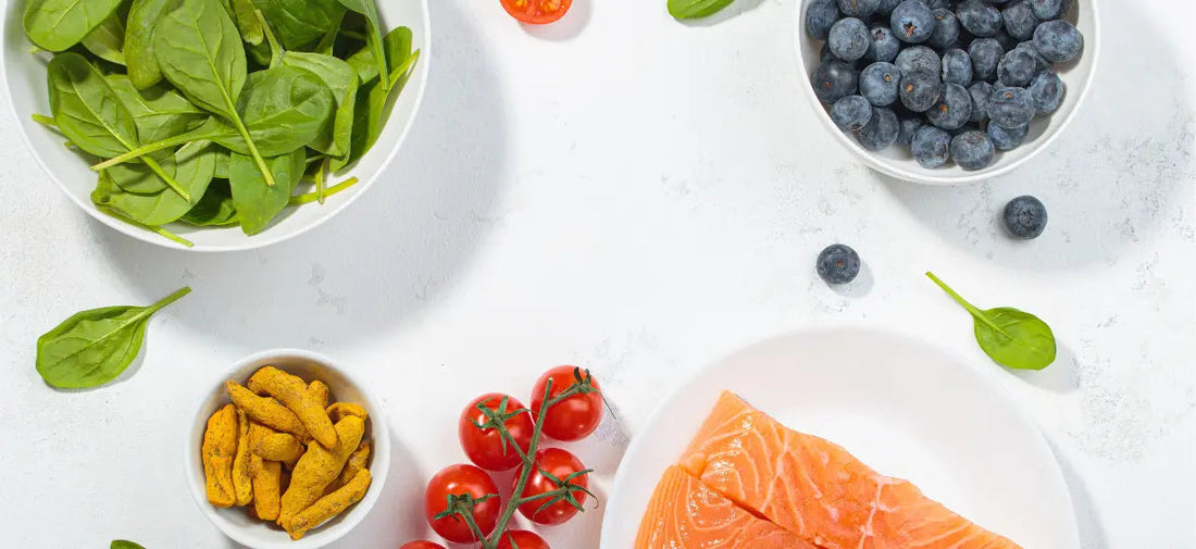 Draufsicht auf einen Tisch mit mehreren Zutaten: oben links eine Schale mit Spinat, oben rechts eine Schale mit Blaubeeren, unten links eine Schale mit Sojaschnetzeln, daneben Strauchtomaten und ein Teller mit rohem Lachsfilet.