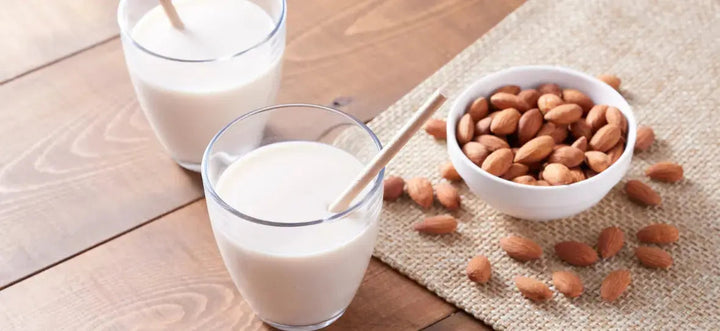 Zwei Gläser mit Mandelmilch und Strohhalmen stehen nebeneinander auf einem Tisch. Daneben befindet sich eine Schüssel mit Mandeln, umgeben von weiteren Mandeln auf einer beigen Tischdecke.