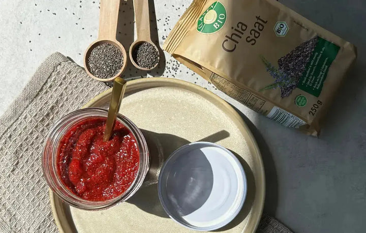 Draufsicht auf ein Glas mit Chia-Marmelade auf einem Teller, der Glasdeckel liegt daneben. Daneben befinden sich eine Packung Chiasamen und zwei Holzlöffel mit losen Chiasamen auf dem Tisch.