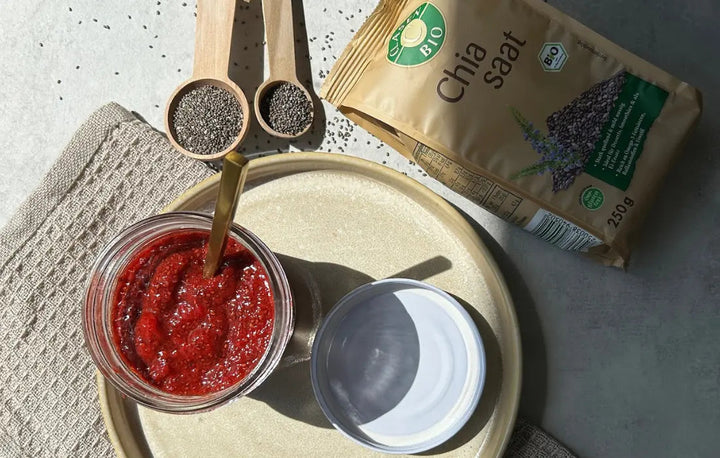 Draufsicht auf ein Glas mit Chia-Marmelade auf einem Teller, der Glasdeckel liegt daneben. Daneben befinden sich eine Packung Chiasamen und zwei Holzlöffel mit losen Chiasamen auf dem Tisch.