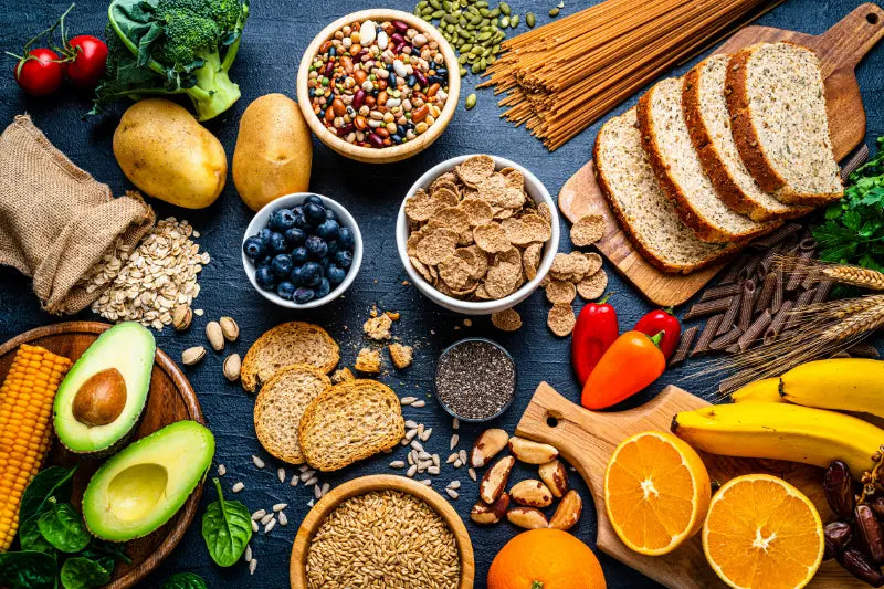 Verschiedene ballaststoffreiche Lebensmittel liegen auf einem Tisch, darunter Vollkornbrot, Haferflocken, Nudeln, Kartoffeln, Hülsenfrüchte, Nüsse, Samen, Avocado, Bananen, Orangen, Blaubeeren, Brokkoli, Mais und Paprika.