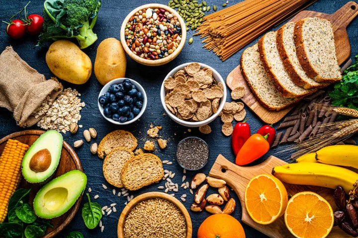Verschiedene ballaststoffreiche Lebensmittel liegen auf einem Tisch, darunter Vollkornbrot, Haferflocken, Nudeln, Kartoffeln, Hülsenfrüchte, Nüsse, Samen, Avocado, Bananen, Orangen, Blaubeeren, Brokkoli, Mais und Paprika.