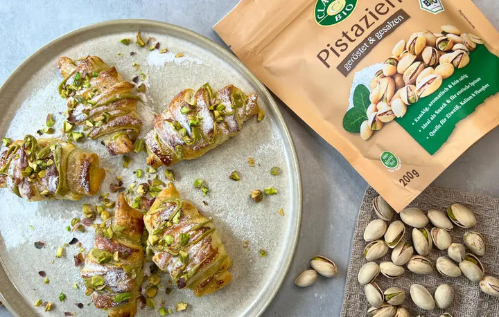 Draufsicht auf einen Teller mit fünf Pistazien-Croissants, daneben eine Tüte Pistazien und lose Pistazien.