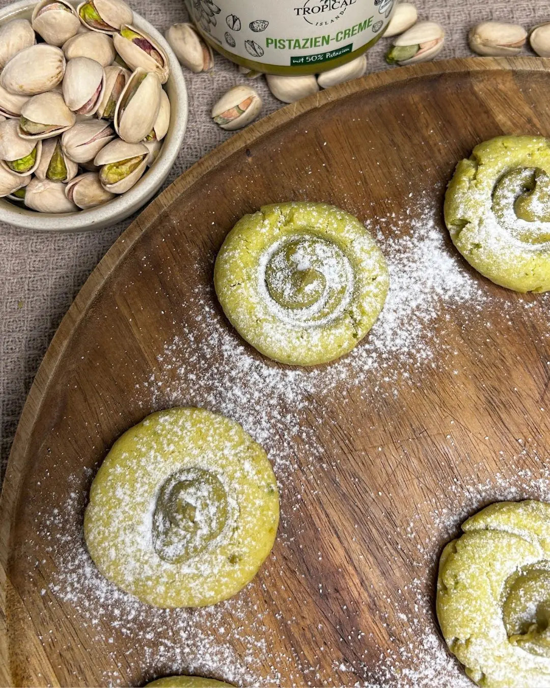 Mehrere runde, hellgrüne Pistazienplätzchen mit einer spiralförmigen Pistaziencreme-Füllung liegen auf einem großen Holzteller und sind mit Puderzucker bestreut. Am oberen Bildrand stehen eine Schale mit Pistazien und ein Glas Pistazien‑Creme.