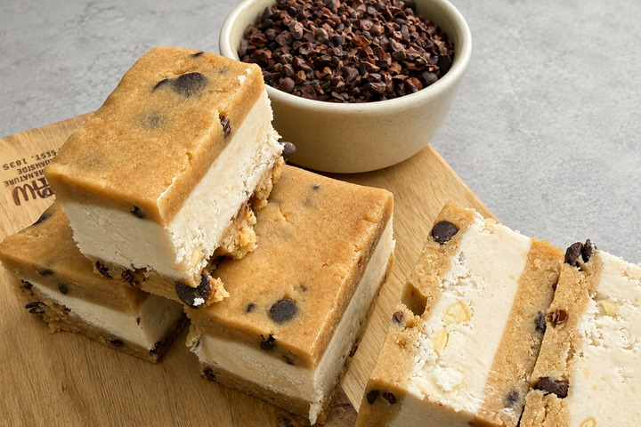 Mehrere vegane Cookie-Dough-Eisriegel mit cremiger Füllung, Schokostückchen und Kakaonibs auf einem Holzbrett, daneben eine Schale mit Kakaonibs.