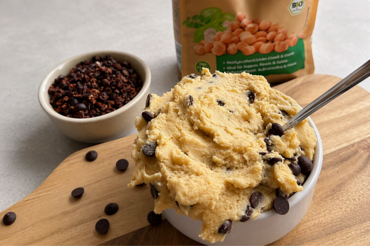 Proteinreicher Kichererbsen-Cookie-Dough mit Schokodrops in einer Schale auf Holzbrett, dekoriert mit Kakaonibs und Kichererbsen im Hintergrund.