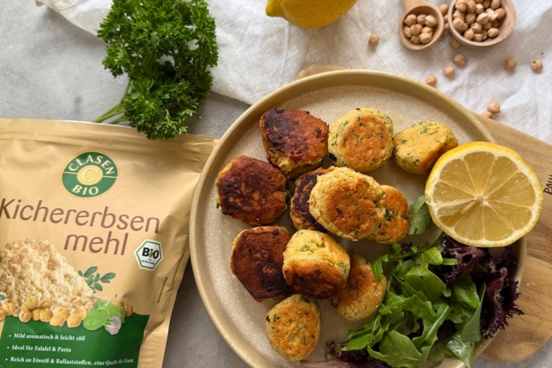 Nahaufnahme eines Tellers mit goldbraunen Kichererbsen-Falafel, frischem Blattsalat und einer halben Zitrone. Daneben steht eine Packung Clasen Bio Kichererbsenmehl, umgeben von Petersilie, Kichererbsen und Küchenutensilien.  
