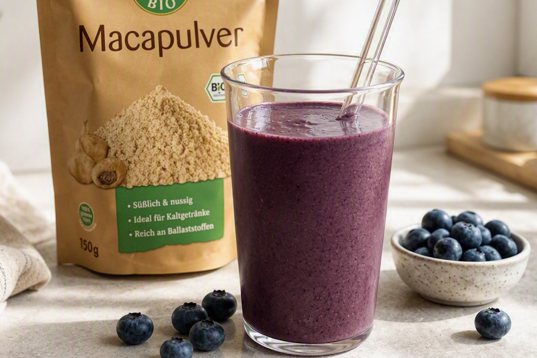Blaubeer-Maca-Smoothie in einem Glas mit Glasstrohhalm, daneben eine Packung Bio-Macapulver auf einer hellen Küchenarbeitsplatte.