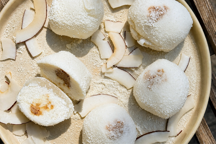 Kokos-Mochis auf einem Teller, teilweise aufgeschnitten, mit sichtbarer cremiger Füllung und Kokoschips.