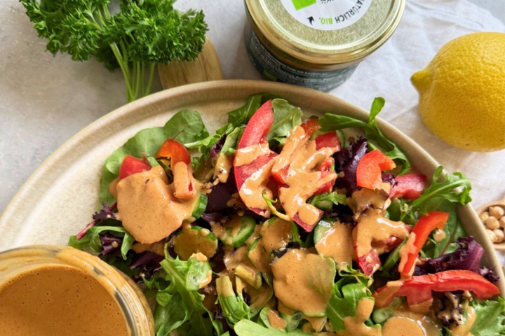 Frischer gemischter Salat mit Blattsalaten, Gurke und roter Paprika in einer Schale, großzügig mit cremigem Dressing beträufelt. Daneben liegen eine Zitrone, frische Petersilie, ein Glas mit Dressing sowie einige Nüsse auf einem hellen Untergrund.