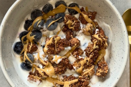 Bowl mit Joghurt, Heidelbeeren, Schoko-Granola und Nussmus-Topping, von oben fotografiert auf hellem Untergrund.