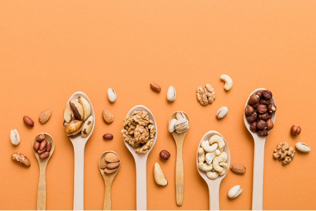 Auswahl verschiedener Nüsse in Holzlöffeln auf orangefarbenem Hintergrund mit Mandeln, Walnüssen, Cashewkernen, Pistazien, Haselnüssen und Paranüssen.