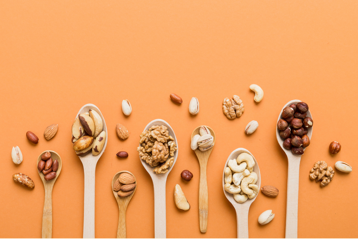 Auswahl verschiedener Nüsse in Holzlöffeln auf orangefarbenem Hintergrund mit Mandeln, Walnüssen, Cashewkernen, Pistazien, Haselnüssen und Paranüssen.