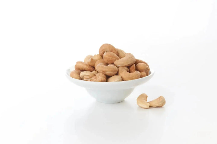 Weiße Schale gefüllt mit naturbelassenen Cashewkernen auf weißem Hintergrund, einige Cashews liegen daneben.