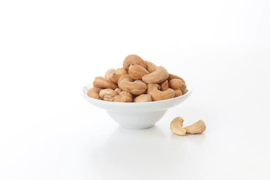 Weiße Schale gefüllt mit naturbelassenen Cashewkernen auf weißem Hintergrund, einige Cashews liegen daneben.