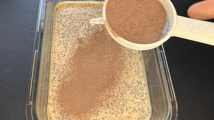 Chiapudding im Tiramisu-Stil in einem Glasbehälter, mit Kakaopulver bestreut, sichtbar beim Bestreuen mit Schokolade