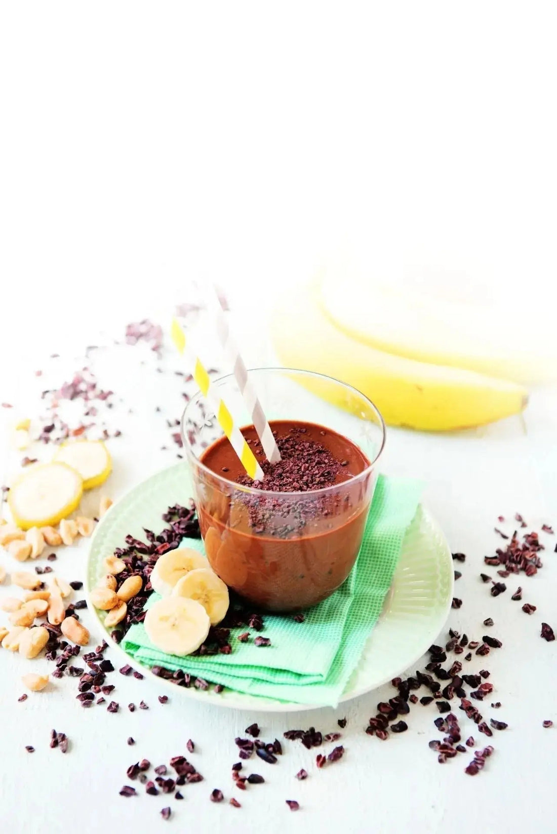 Cremiger Schoko-Erdnuss-Smoothie in einem Glas, bestreut mit Kakaonibs, daneben Erdnüsse und Kakaonibs als Dekoration