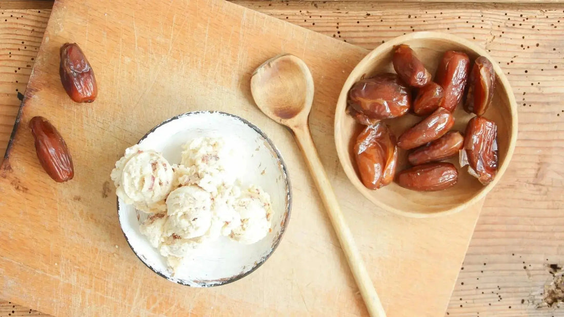 Von oben fotografierter Holztisch mit einem Holzbrett, darauf eine kleine Holzschale mit getrockneten Datteln und ein Holzlöffel. Daneben eine weiße Schale mit weißem Eis und Dattelstückchen, daneben zwei weitere getrocknete Datteln.