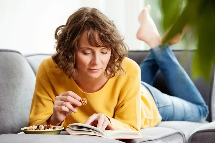 Frau liegt entspannt auf einem grauen Sofa, liest ein Buch und isst Nüsse. Sie trägt einen gelben Pullover und blaue Jeans.