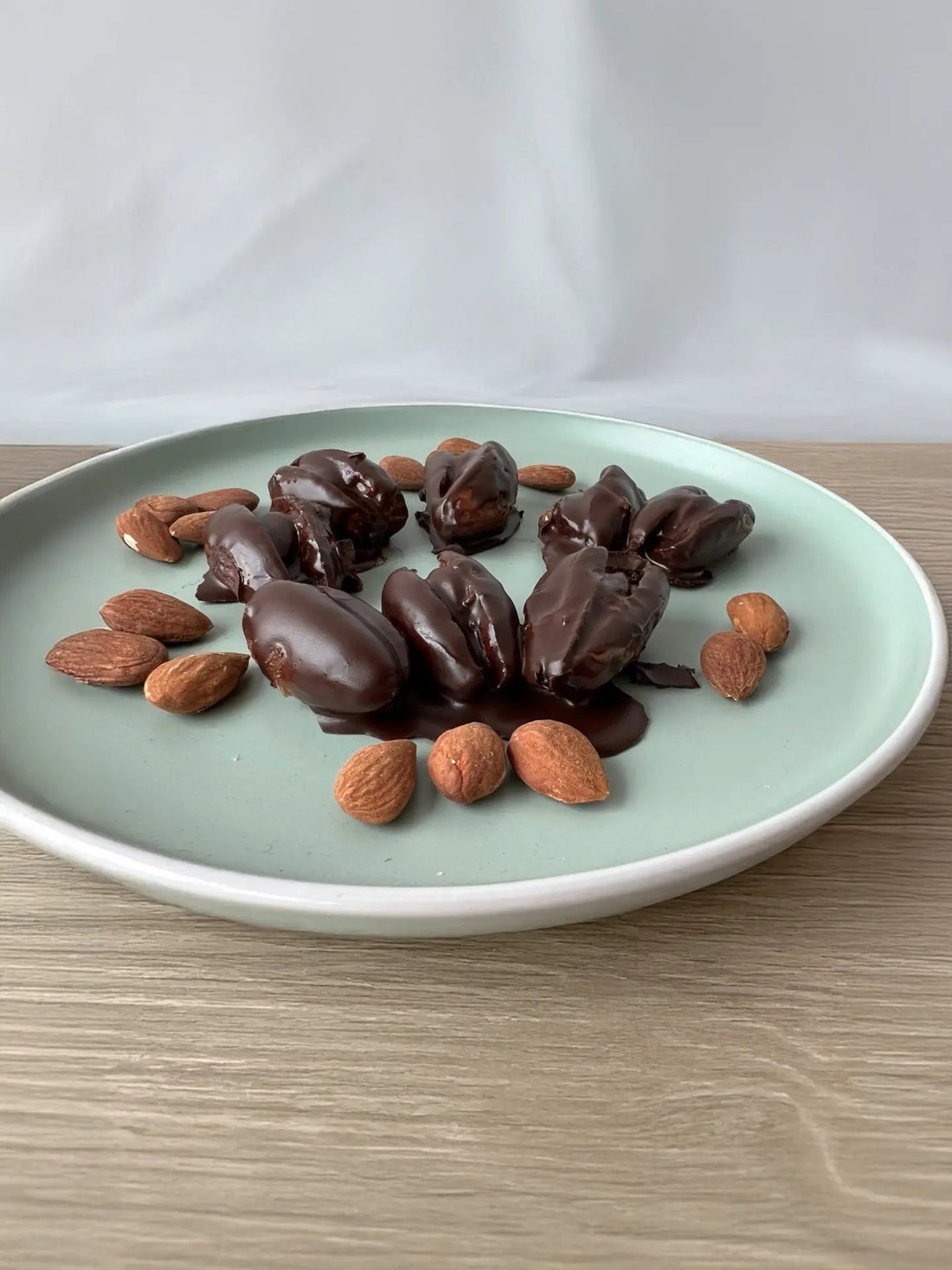 Gefüllte Schokodatteln mit Mandeln und Nussmus, serviert auf einem blau-grünen Porzellanteller, daneben ganze Mandeln als Dekoration