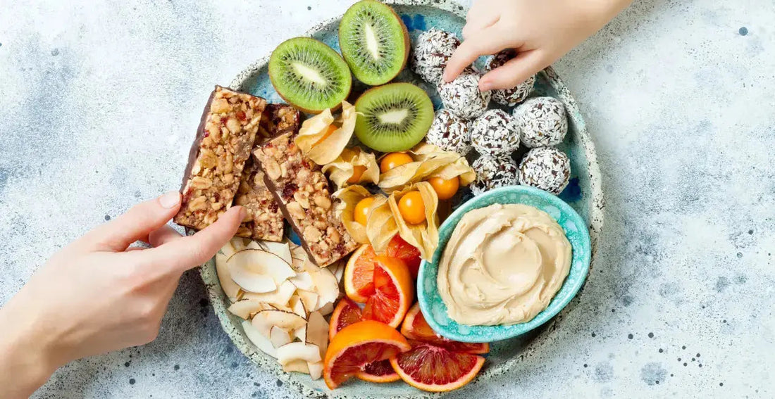 Von oben fotografierter Teller mit verschiedenen gesunden Snacks wie Kokoschips, Müsliriegeln, getrockneter Physalis, Kiwi, Energyballs und einem Dip. Eine Hand greift gerade nach einem Müsliriegel.