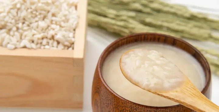 Holzschale mit ungekochtem Getreide, daneben eine kleine Schüssel mit fermentiertem Getreide. Ein Löffel nimmt gerade etwas von dem fermentierten Getreide heraus.