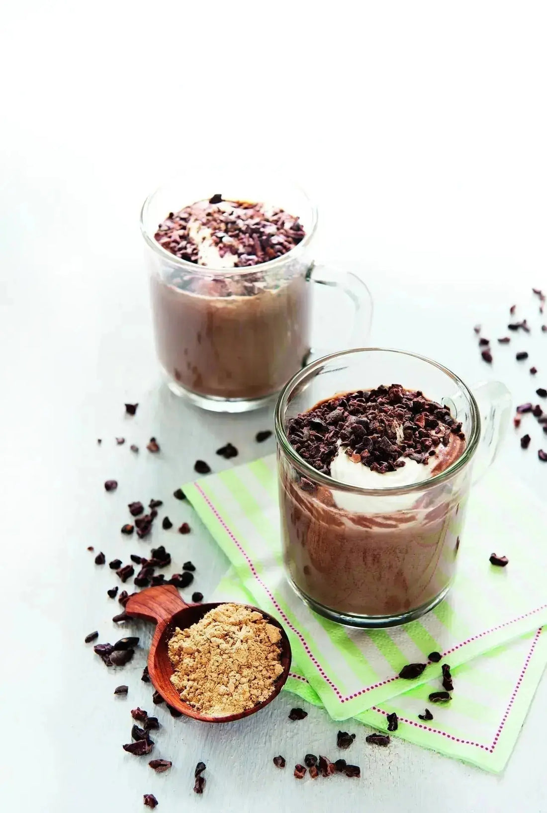 Zwei Gläser Happy Chocolate mit Macapulver, daneben ein Löffel Macapulver und Kakaonibs