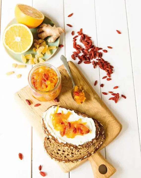 Karotten-Orangen-Marmelade mit Ingwer und Gojibeeren auf Brot mit weißem Käse, serviert auf einem Holzbrett mit frischen Orangenscheiben und Ingwerstücken