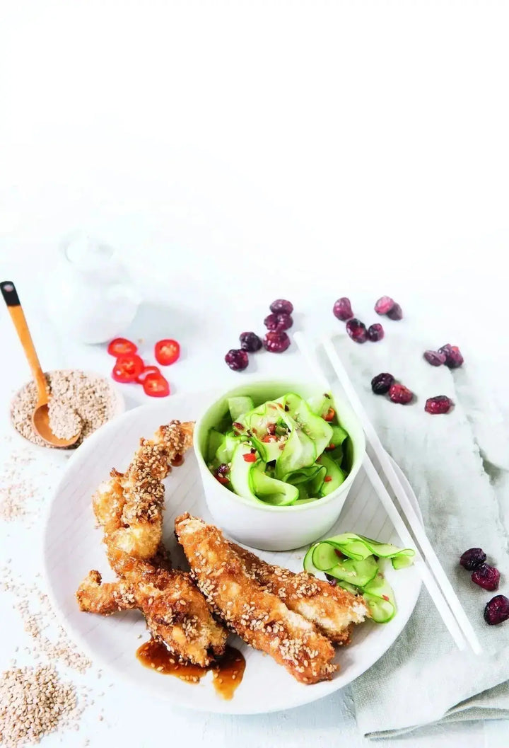 Knuspriges Honig-Sesam-Hühnchen auf einem weißen Teller, serviert mit Gurken-Cranberry-Salat und dekorativen Kräutern