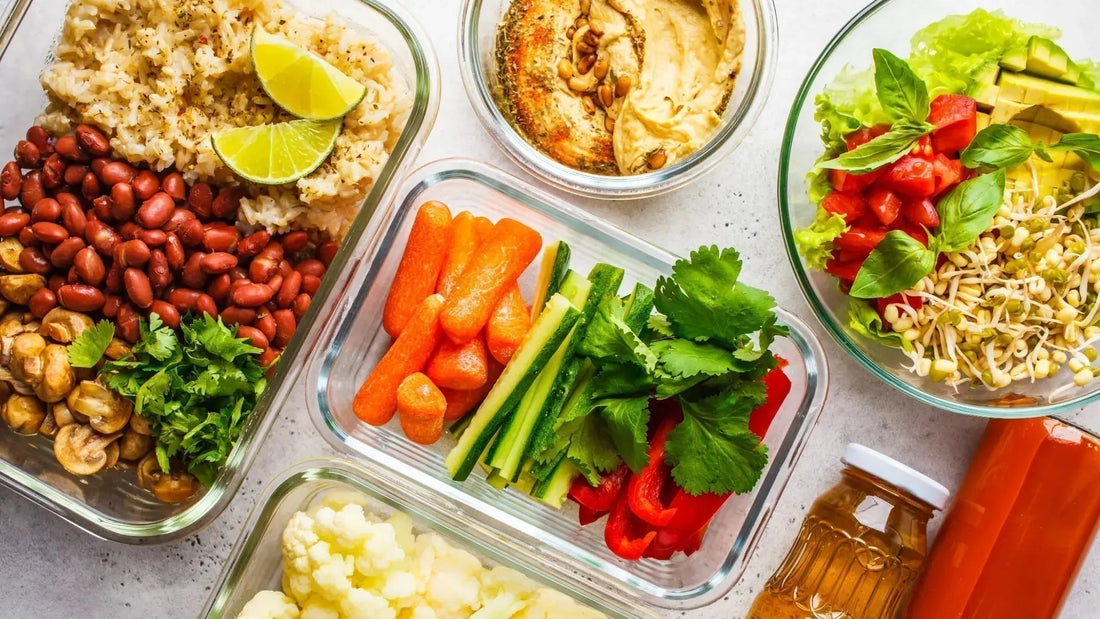Von oben fotografierter hellgrauer Tisch mit mehreren durchsichtigen Glas-Meal-Prep-Behältern. In den Behältern befinden sich verschiedene vorbereitete Gerichte wie Bohnen mit Reis, gedämpftes Gemüse und weitere ausgewogene Mahlzeiten.