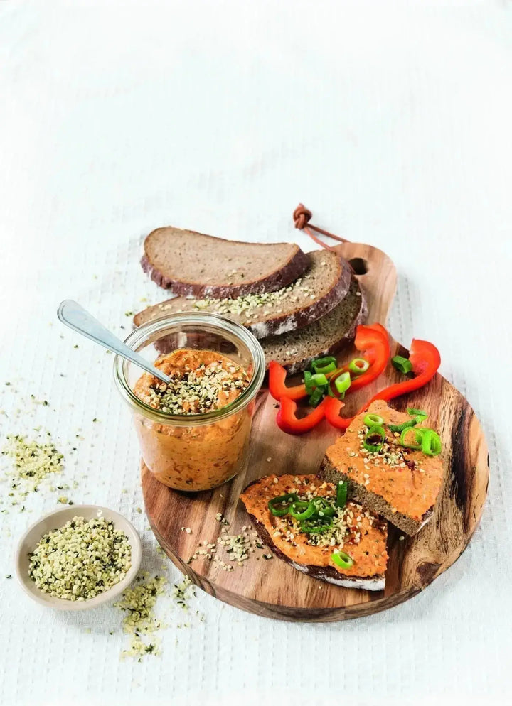 „Mediterraner Hanfsamen-Aufstrich in einem Glas, serviert mit Brot, garniert mit Frühlingszwiebeln und Paprika, daneben eine kleine Schale mit Hanfsamen auf einem Holzbrett