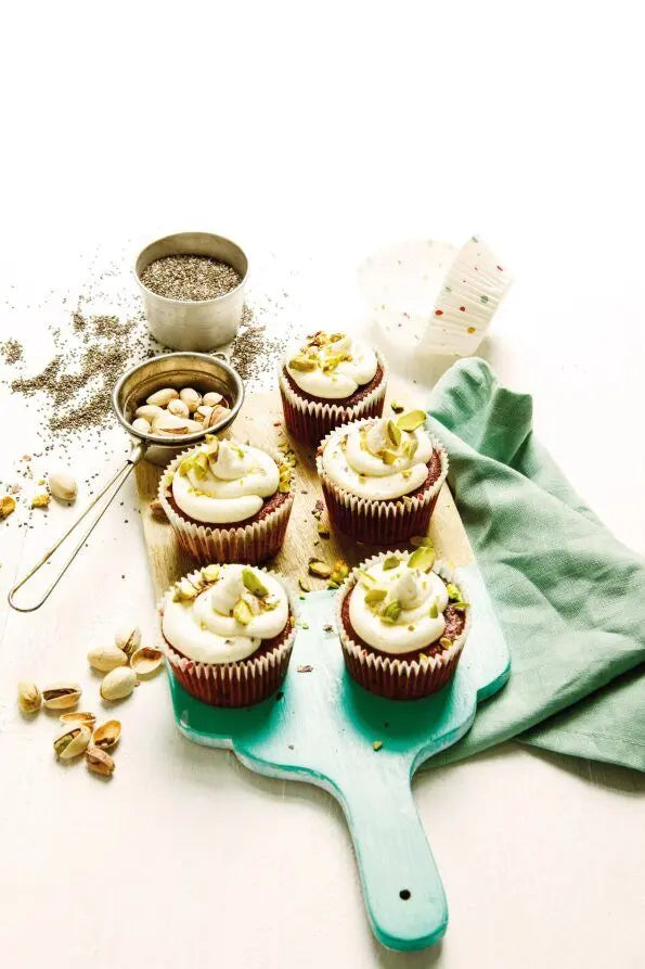 Red Velvet Cupcakes mit Vanille-Frosting und gehackten Pistazien, angerichtet auf einem Holzbrett, daneben Pistazien und Chiasamen als Dekoration