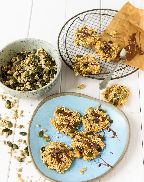 Schoko-Knuspertaler mit Saaten und Salted Caramel auf einem blauen Keramikteller, daneben ein Löffel mit geschmolzener Schokolade und einige Samen