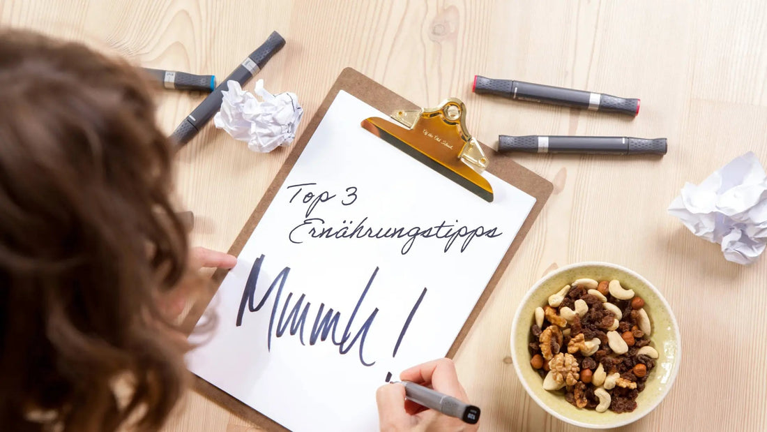 Klemmbrett mit weißem Blatt auf einem Tisch von oben fotografiert. Auf dem Blatt steht „Top 3 Ernährungstipps“, eine Frau schreibt gerade „Mmmh!“ darunter. Daneben liegen vier Stifte, zerknülltes Papier und eine Schüssel mit Studentenfutter.