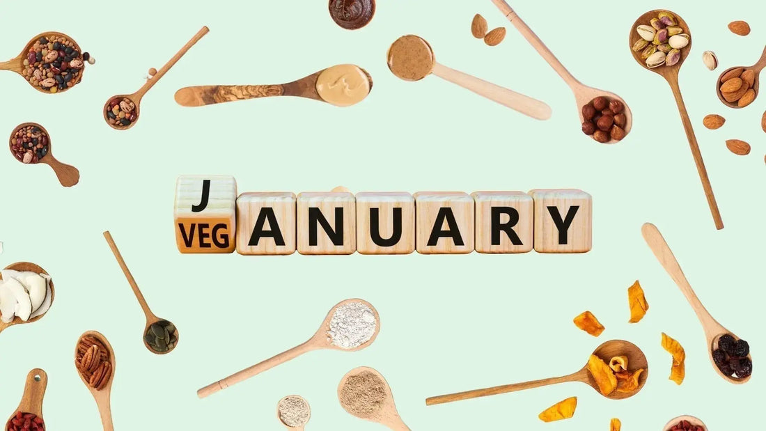 Holzwürfel mit dem Schriftzug „Veganuary“ auf mintfarbenem Hintergrund. Im Hintergrund liegen Holzlöffel mit verschiedenen veganen Zutaten wie Mehl, Hülsenfrüchten, Nüssen, Saaten, Trockenfrüchten und Nussmus.