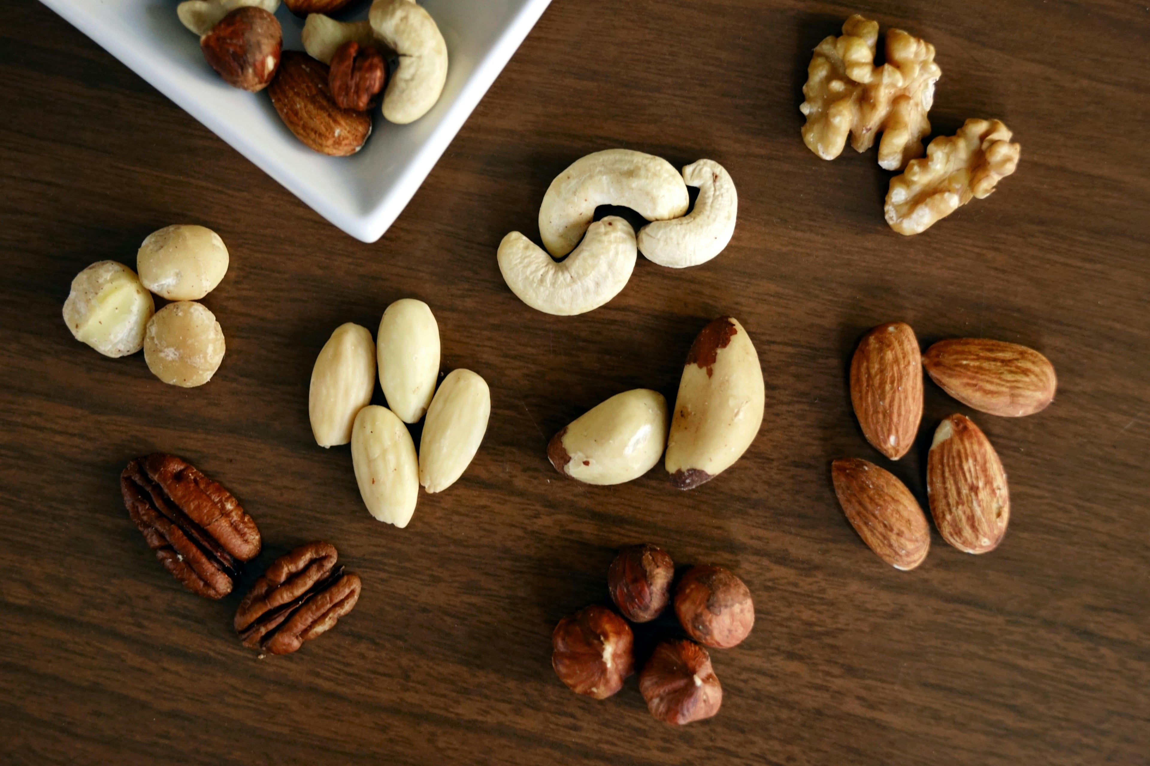 Verschiedene Nussarten auf einem Holztisch, darunter Cashews, Mandeln, Haselnüsse, Pekannüsse, Macadamias, Walnüsse, Paranüsse und Mandeln.