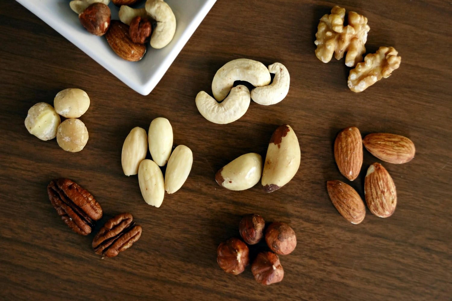Verschiedene Nussarten auf einem Holztisch, darunter Cashews, Mandeln, Haselnüsse, Pekannüsse, Macadamias, Walnüsse, Paranüsse und Mandeln.