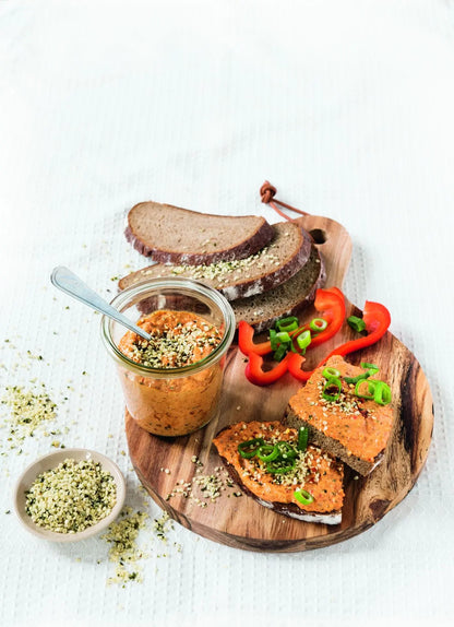 Mediterraner Hanfsamen-Aufstrich in einem Glas, dazu Brot mit Aufstrich, garniert mit Frühlingszwiebeln und Paprika, daneben eine kleine Schale mit Hanfsamen, alles auf einem Holzbrett angerichtet.