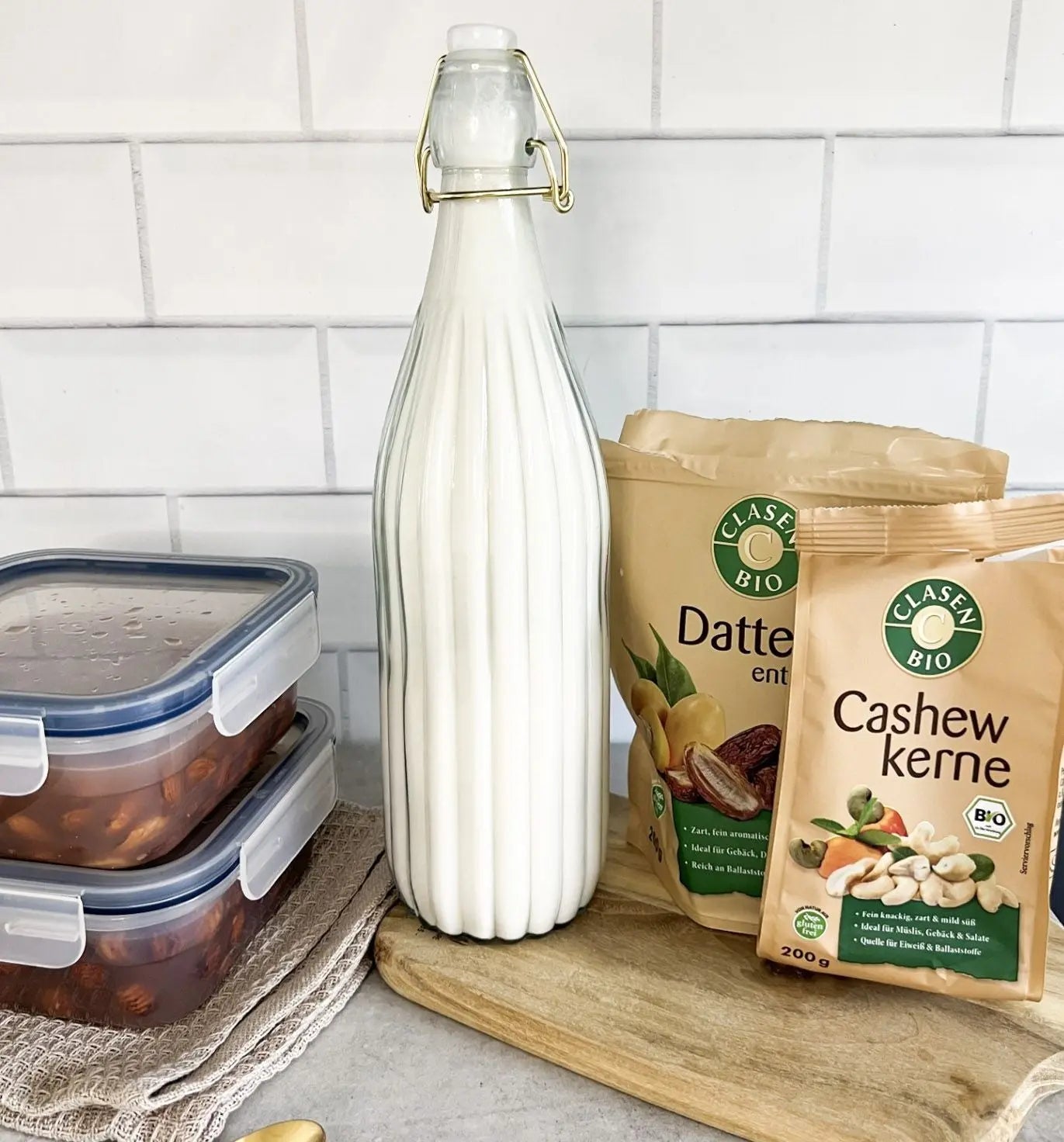Hausgemachte Nussmilch aus Mandeln, Cashews und Haselnüssen in einem Glasbehälter auf einem Holzbrett, daneben Cashewkerne, eine Datteln-Verpackung und zwei Tupperdosen mit über Nacht eingeweichten Nüssen in Wasser.
